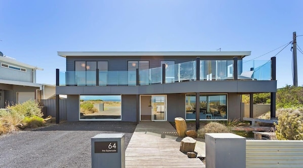 A modern two-story house with a balcony, a driveway, and a number '64' sign. - Ultimate Beachfront Retreat (Sunderland Bay)