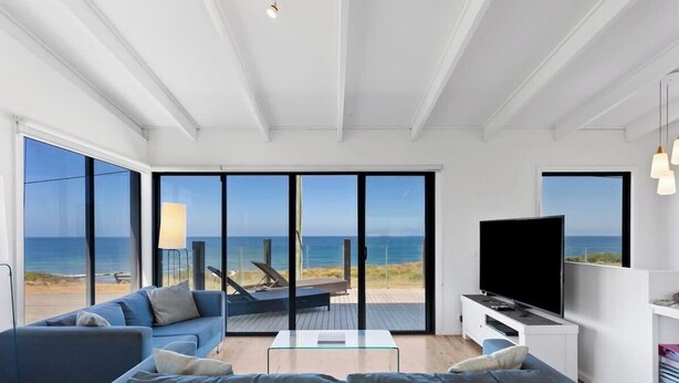 A modern living room with a large TV, a sofa, and a coffee table. There are large windows offering a view of the beach and ocean. - Ultimate Beachfront Retreat (Sunderland Bay)