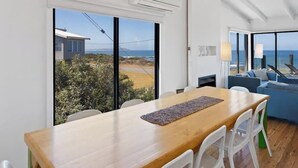 A modern dining area with a wooden table and white chairs, large windows with a view of the sea and a building, and a blue sofa in the background. - Ultimate Beachfront Retreat (Sunderland Bay)