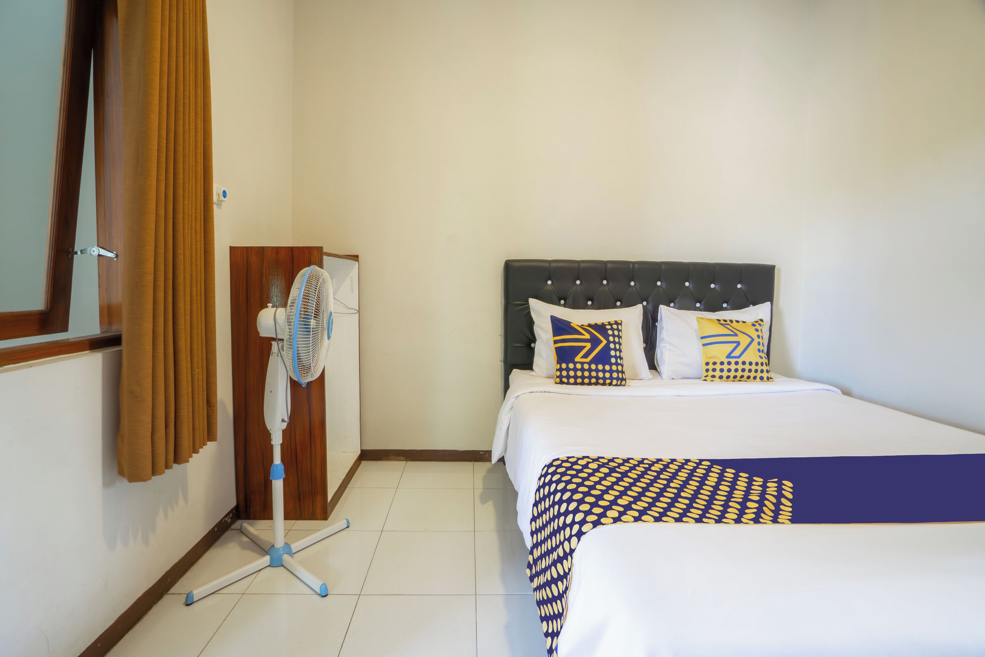 A hotel room with a bed, a standing fan, and a wooden door with curtains.