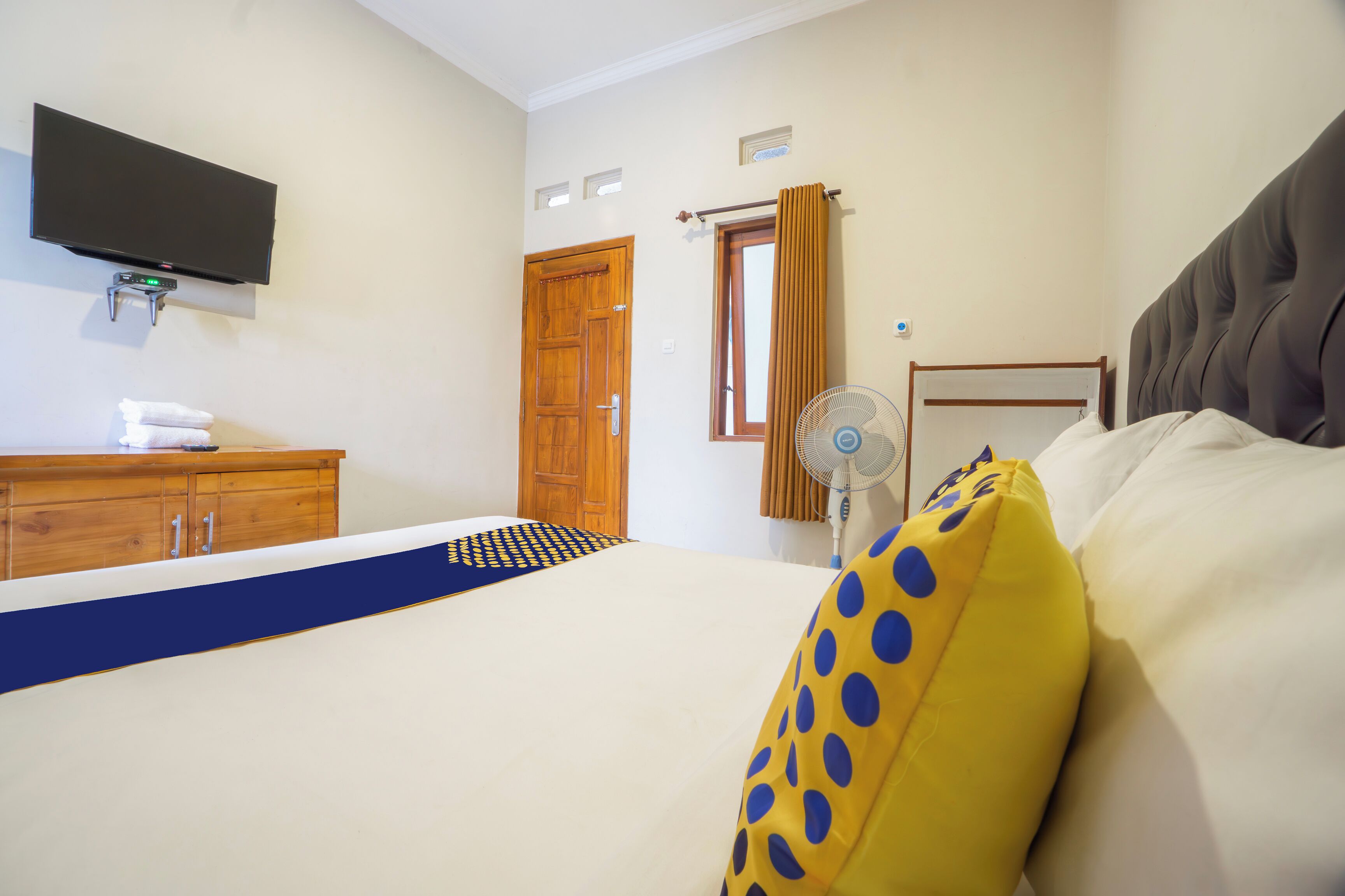 A hotel room with a bed, a TV mounted on the wall, a wooden wardrobe, a window with curtains, and a fan.