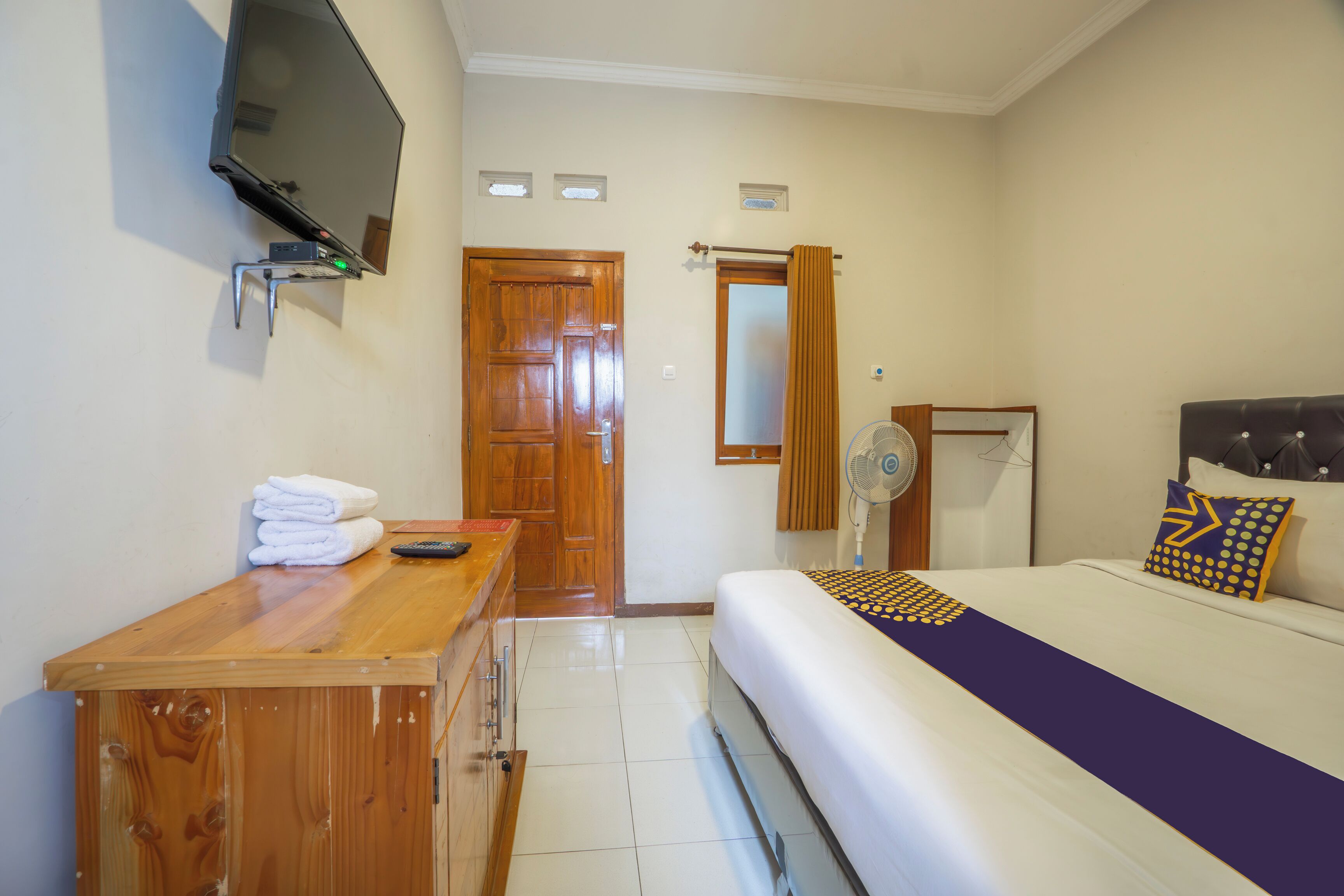 A hotel room with a bed, a wooden desk, a television mounted on the wall, and a door.