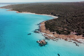 A coastal landscape with clear turquoise waters, a rocky shoreline, and dense forested hills.