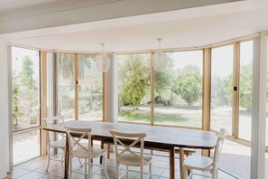 A sunlit room with a large table and chairs, large windows with a view of greenery, and a ceiling light fixture. - Seascape - Where Coastal Fun and Adventure Meet (Eagle Bay)