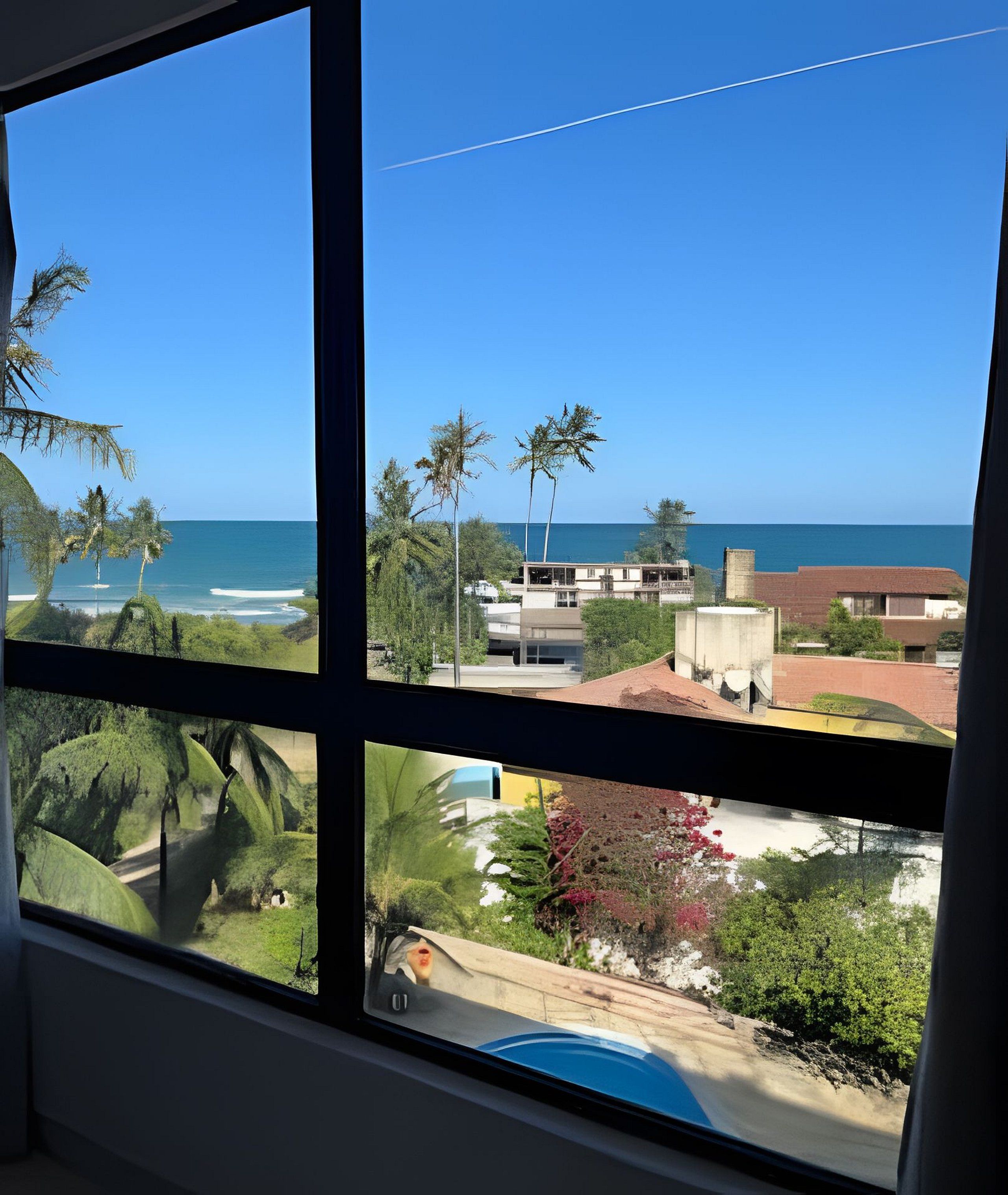 Blick durch ein Fenster auf eine Küstenlandschaft mit Palmen, einem Strand und Gebäuden.