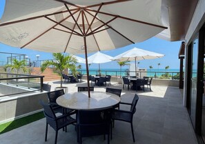 An outdoor patio with round tables and chairs, large umbrellas, and a view of the ocean.