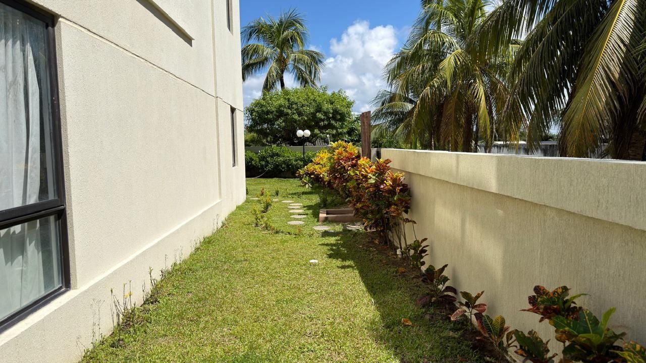 Un sendero estrecho que lleva a un edificio de fachada blanca, rodeado por un césped cuidado y follaje tropical.