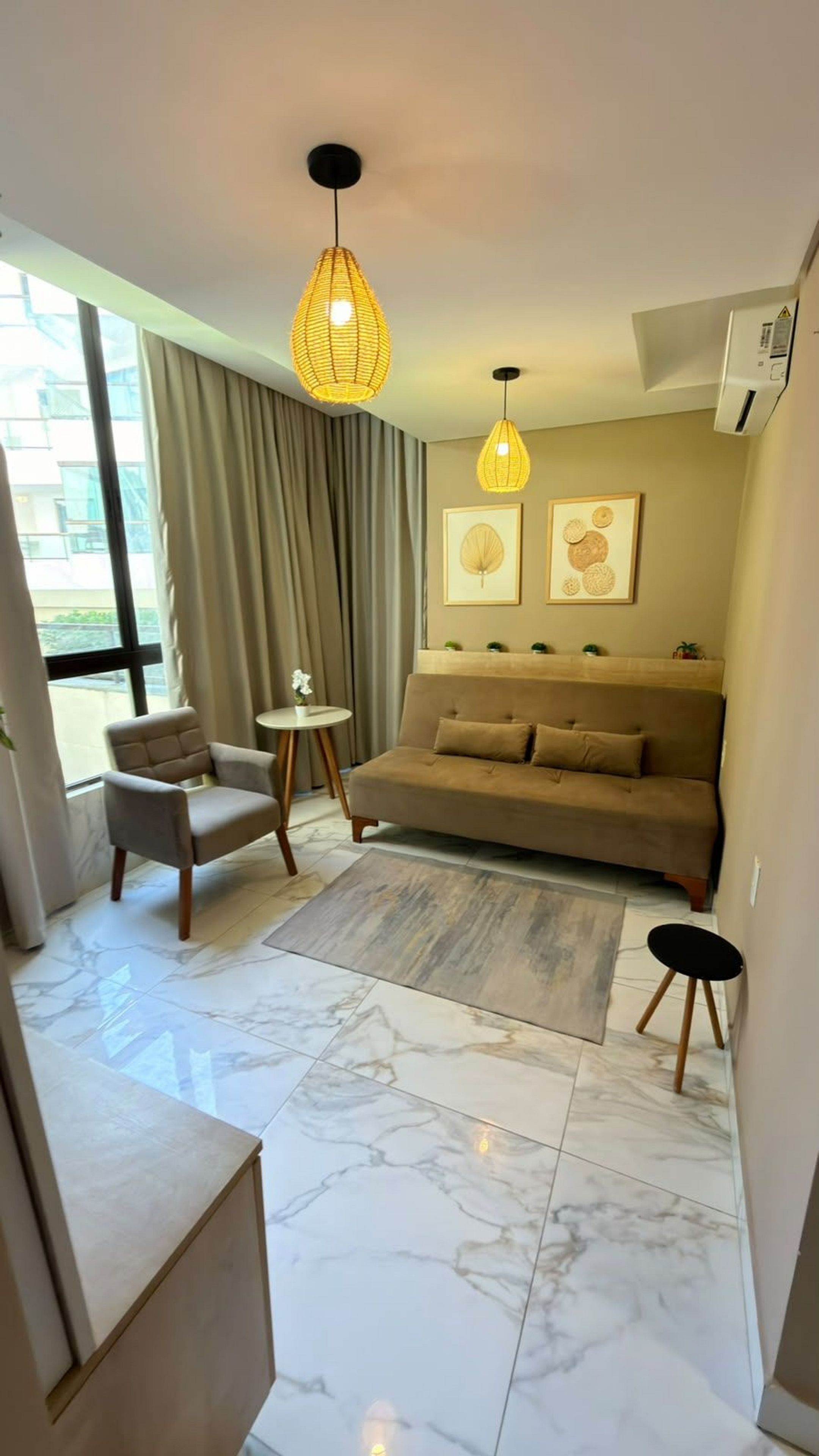 Ein modernes Wohnzimmer mit einer Couch, einem Sessel und einem kleinen Tisch. Es gibt zwei Pendelleuchten, ein Fenster mit Vorhängen und eine Wandklimaanlage.