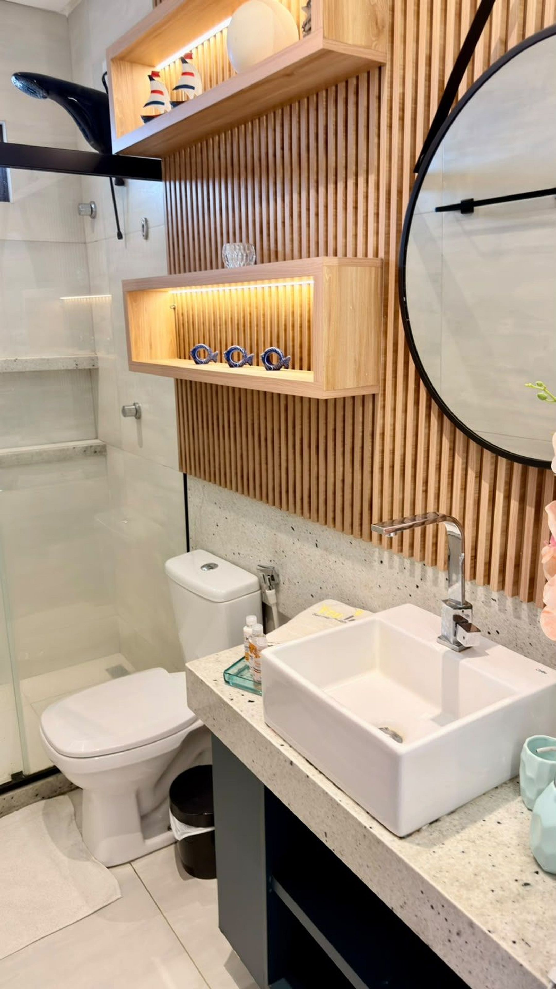 Moderni kylpyhuone, jossa on wc, pesuallas ja suihku. Siellä on puisia hyllyjä, joilla on koriste-esineitä, sekä pyöreä peili.