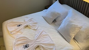 A neatly made bed with white linens and two towels, a window with curtains, and a wall-mounted artwork.