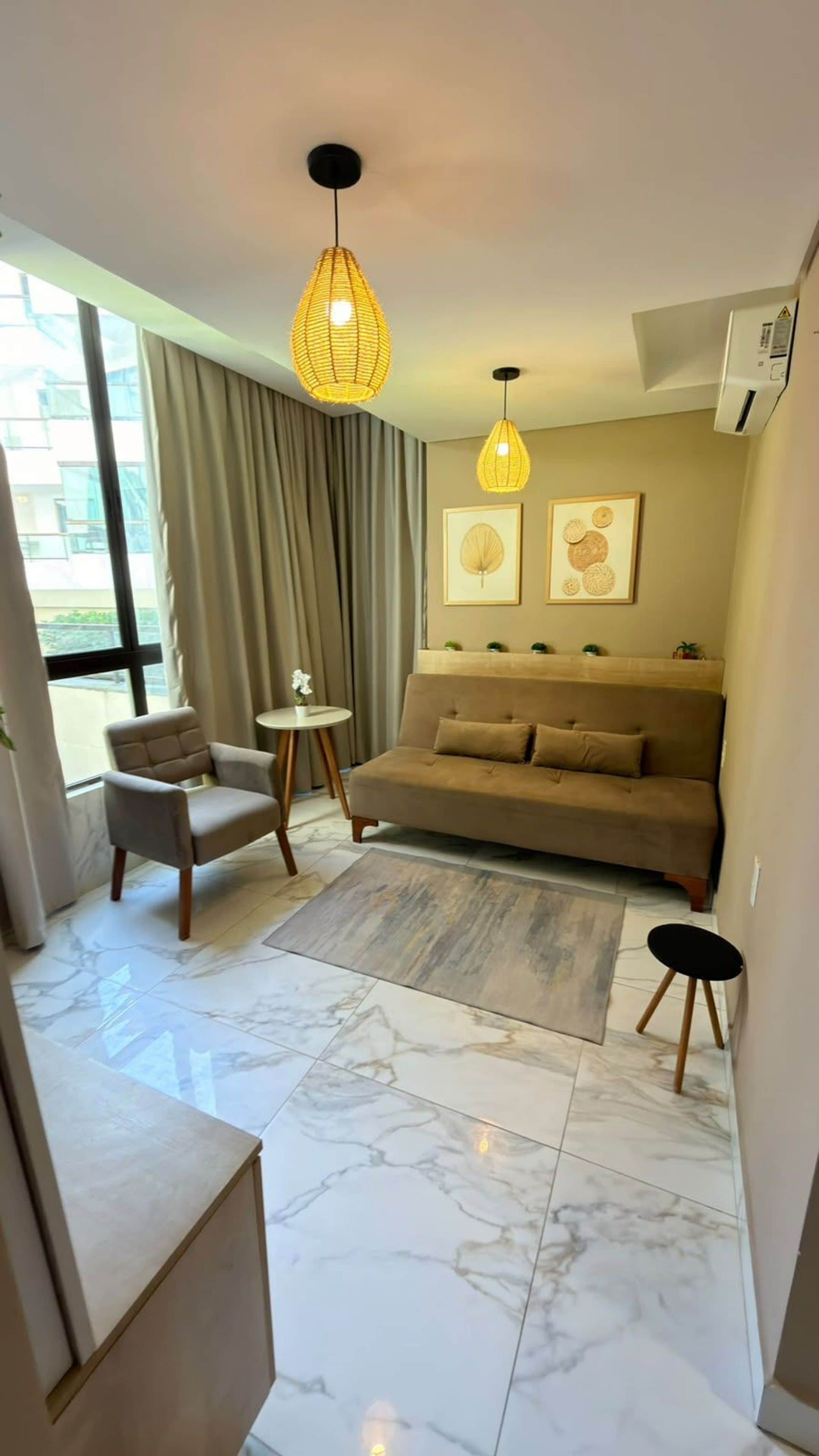 Una sala moderna con sofá, sillón y una mesita. Se observan dos lámparas colgantes, un ventanal con cortinas y un aire acondicionado de pared.
