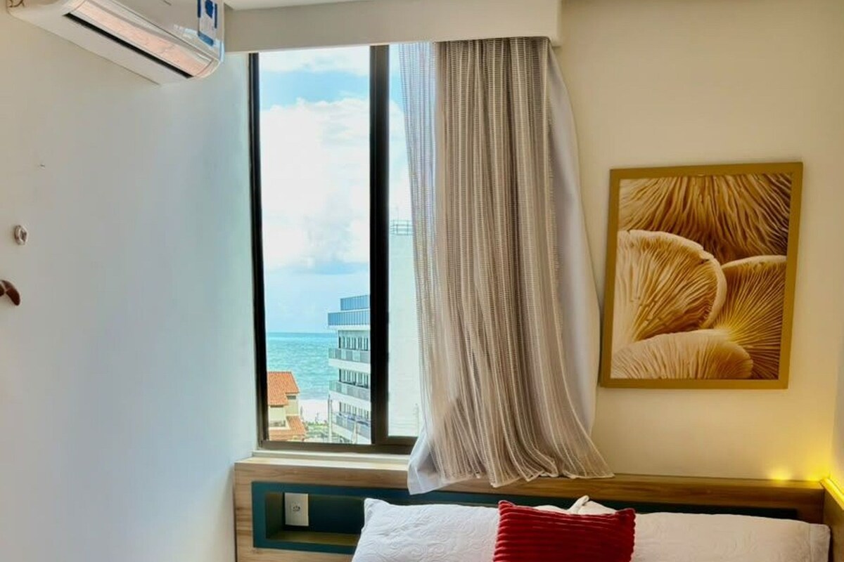 A hotel room with a bed, a window with curtains, a wall-mounted air conditioner, and a painting.