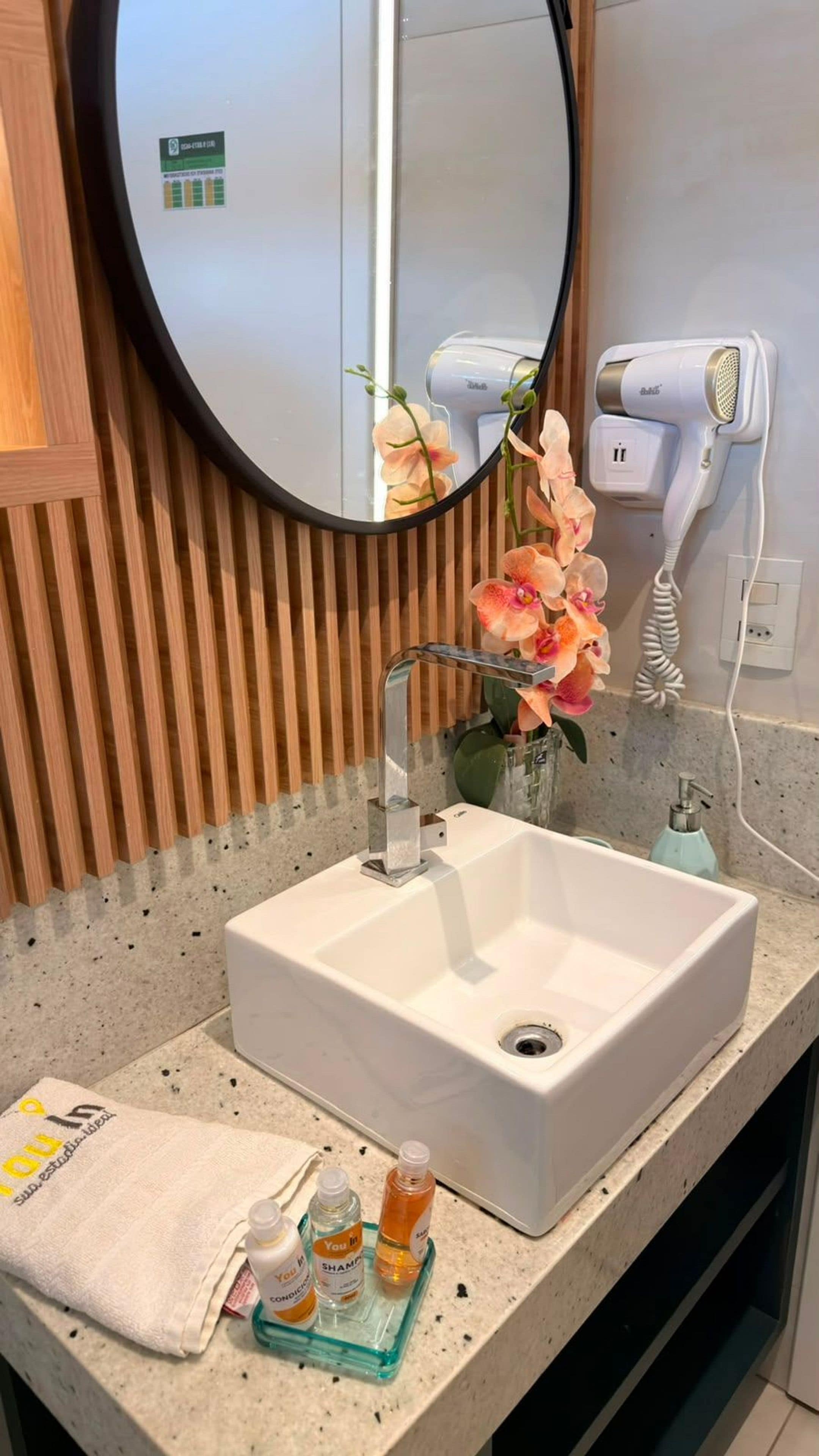 Ein Badezimmer mit weißem Waschbecken, einem runden Spiegel, einem Fön und einem Handtuch auf der Ablagefläche.