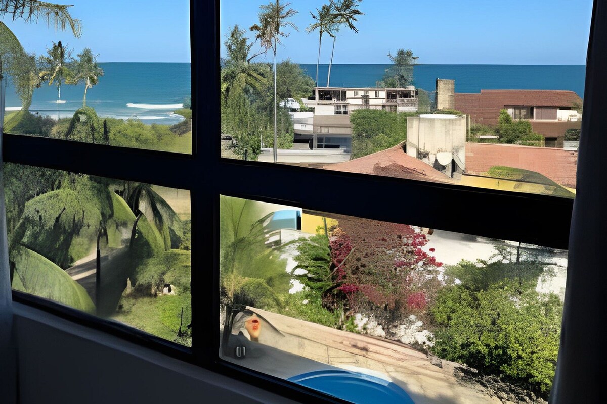A view through a window of a coastal area with palm trees, a beach, and buildings.