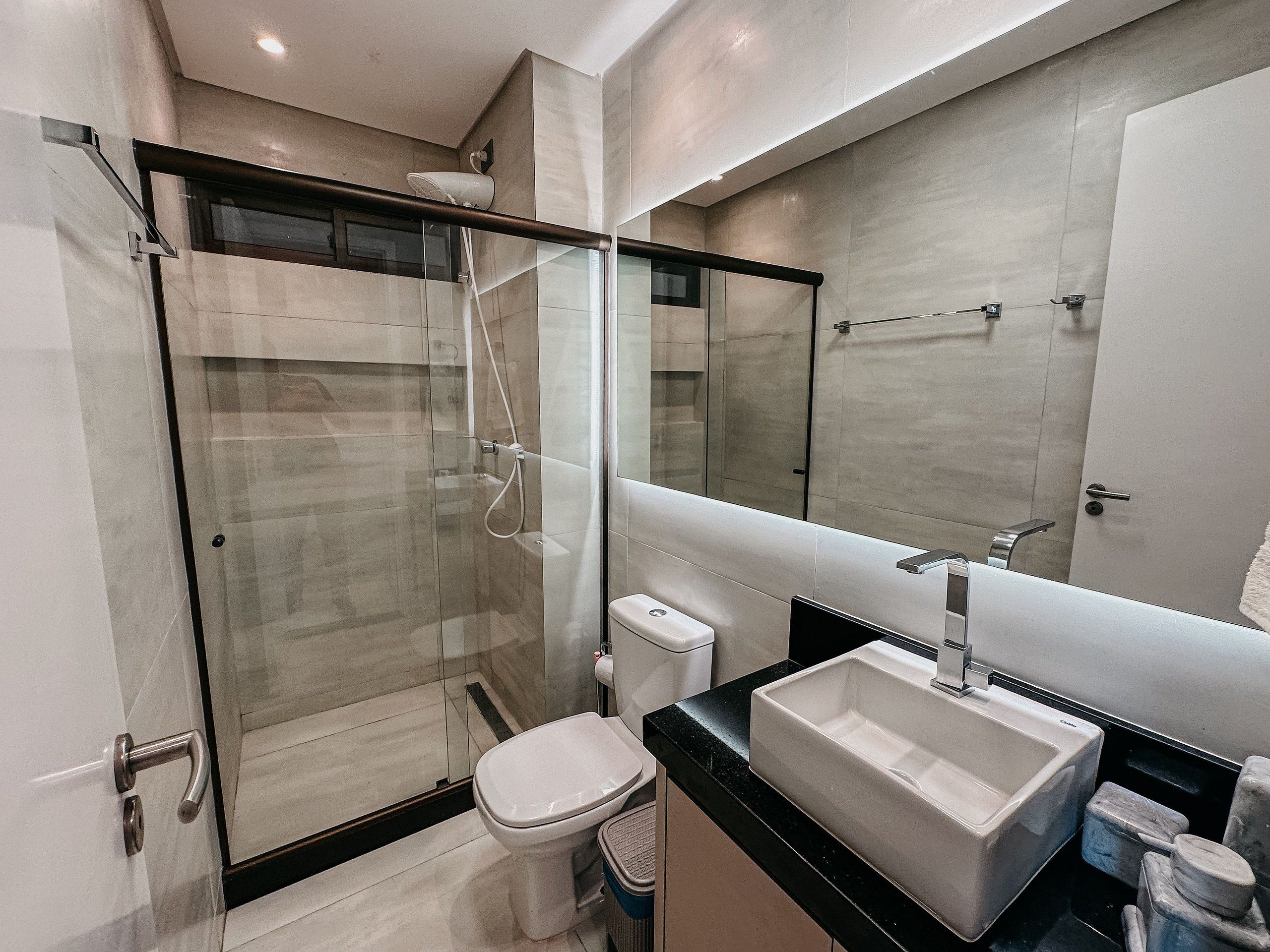 Uma casa de banho moderna com chuveiro separado por vidro, sanita branca e espelho grande sobre bancada preta com lavatório branco.