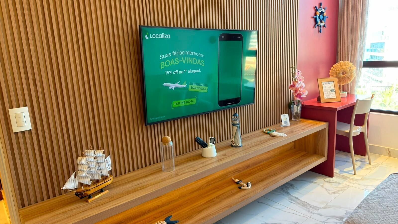 Zona de recepción de un hotel con un televisor mostrando un mensaje promocional, un mostrador de madera y una silla.