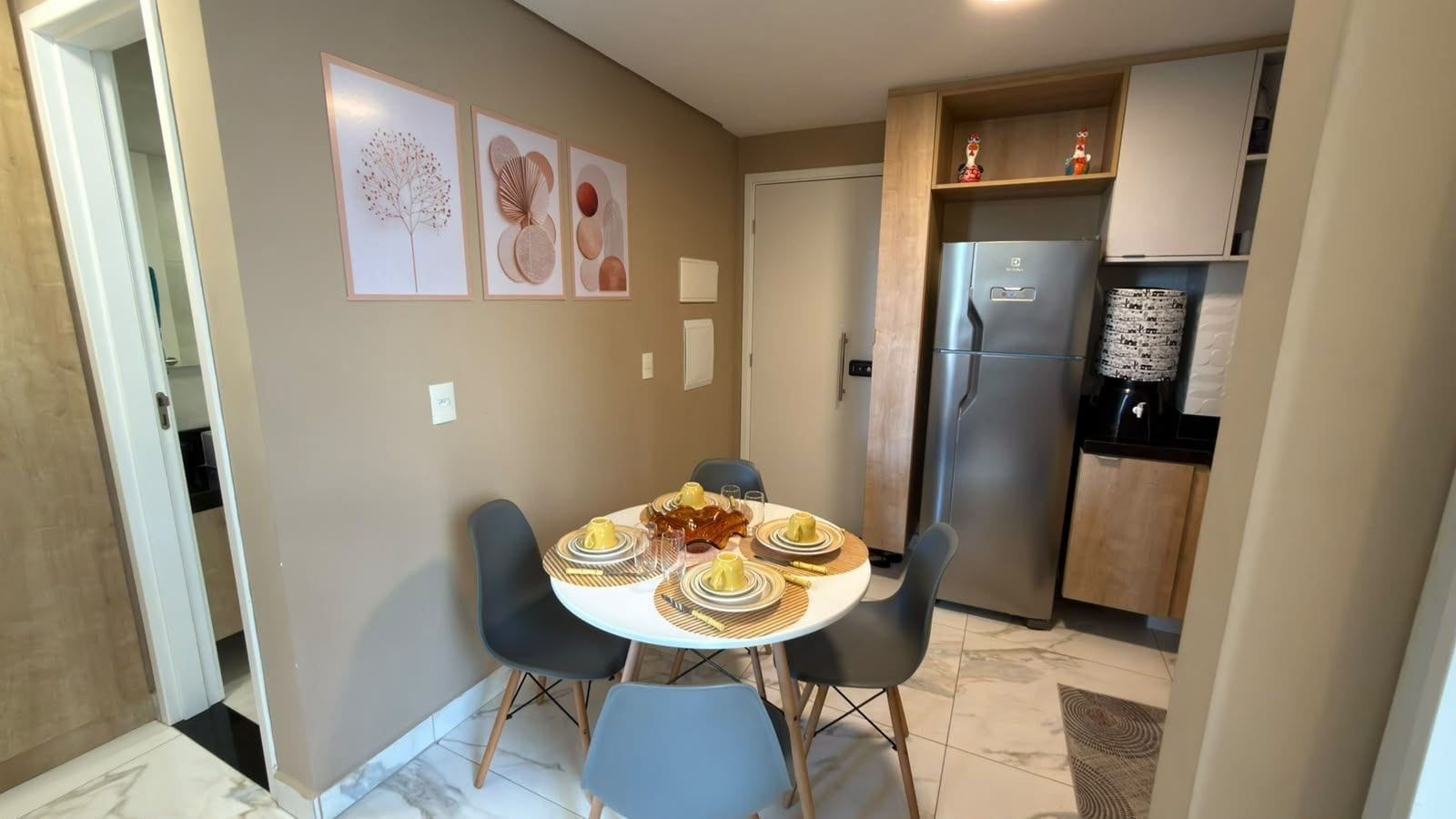 A dining area with a round table set for four, surrounded by chairs. A refrigerator and cabinets are visible in the background.