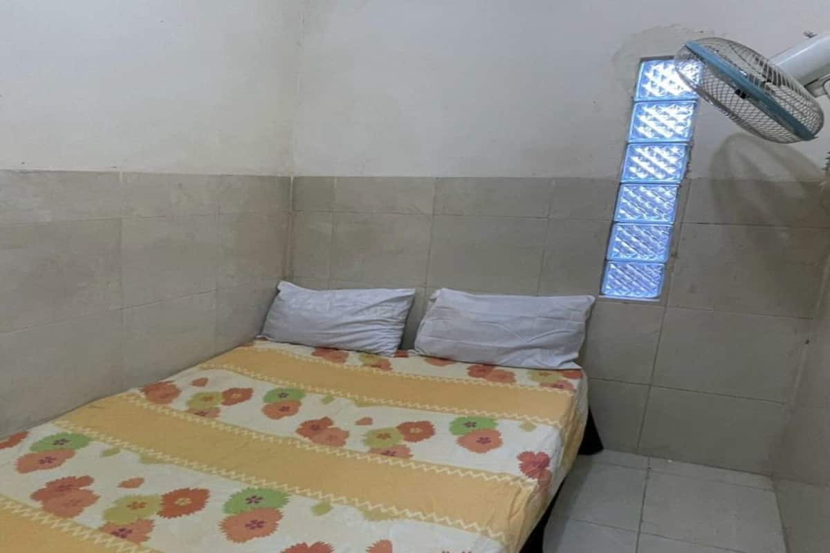 Habitació petita amb un llit individual, un edredó amb motius florals, un ventilador de peu i una finestra amb reixeta metàl·lica.