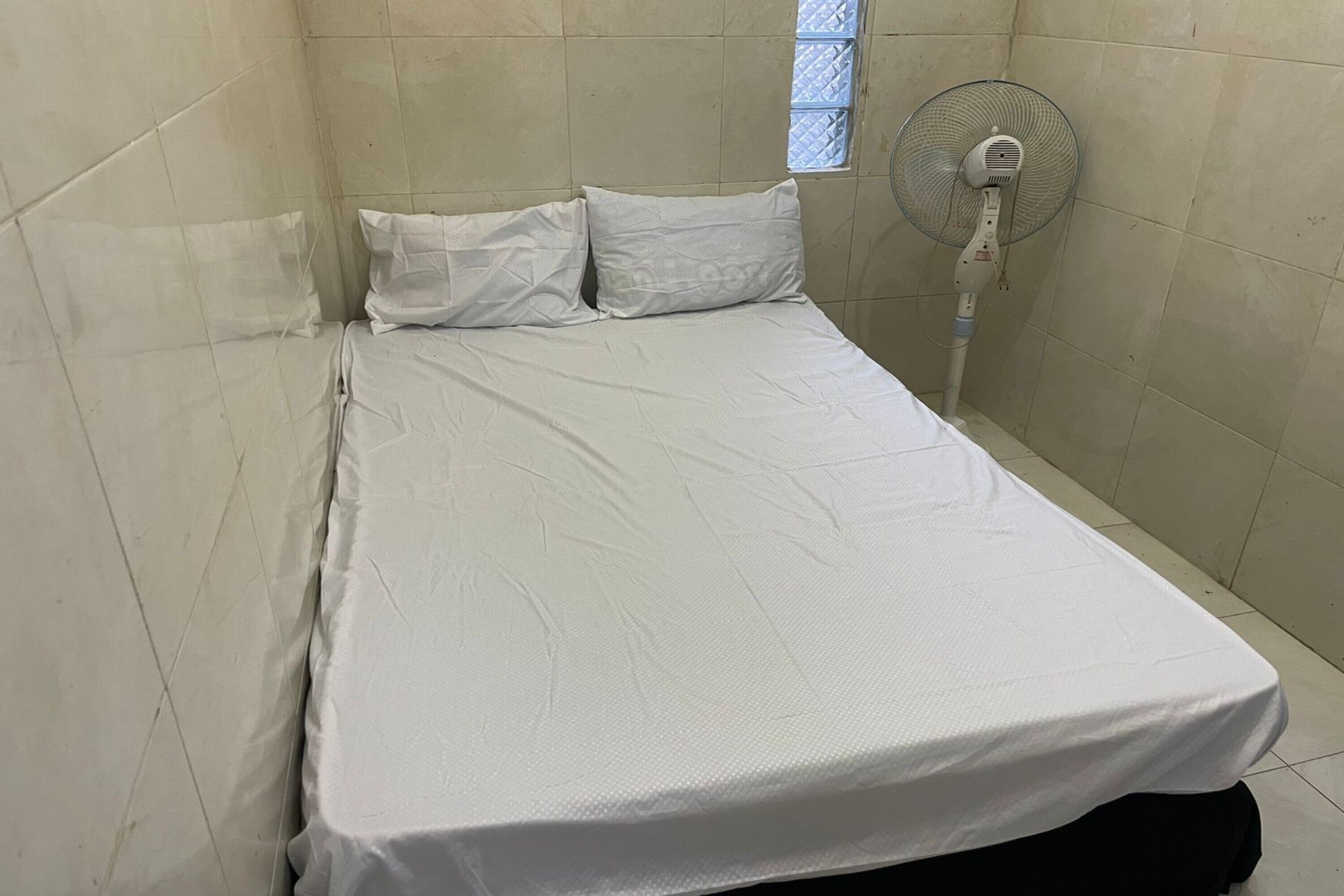 A small, single-bed room with a white mattress and pillows, a standing fan, and a window with a grid pattern.