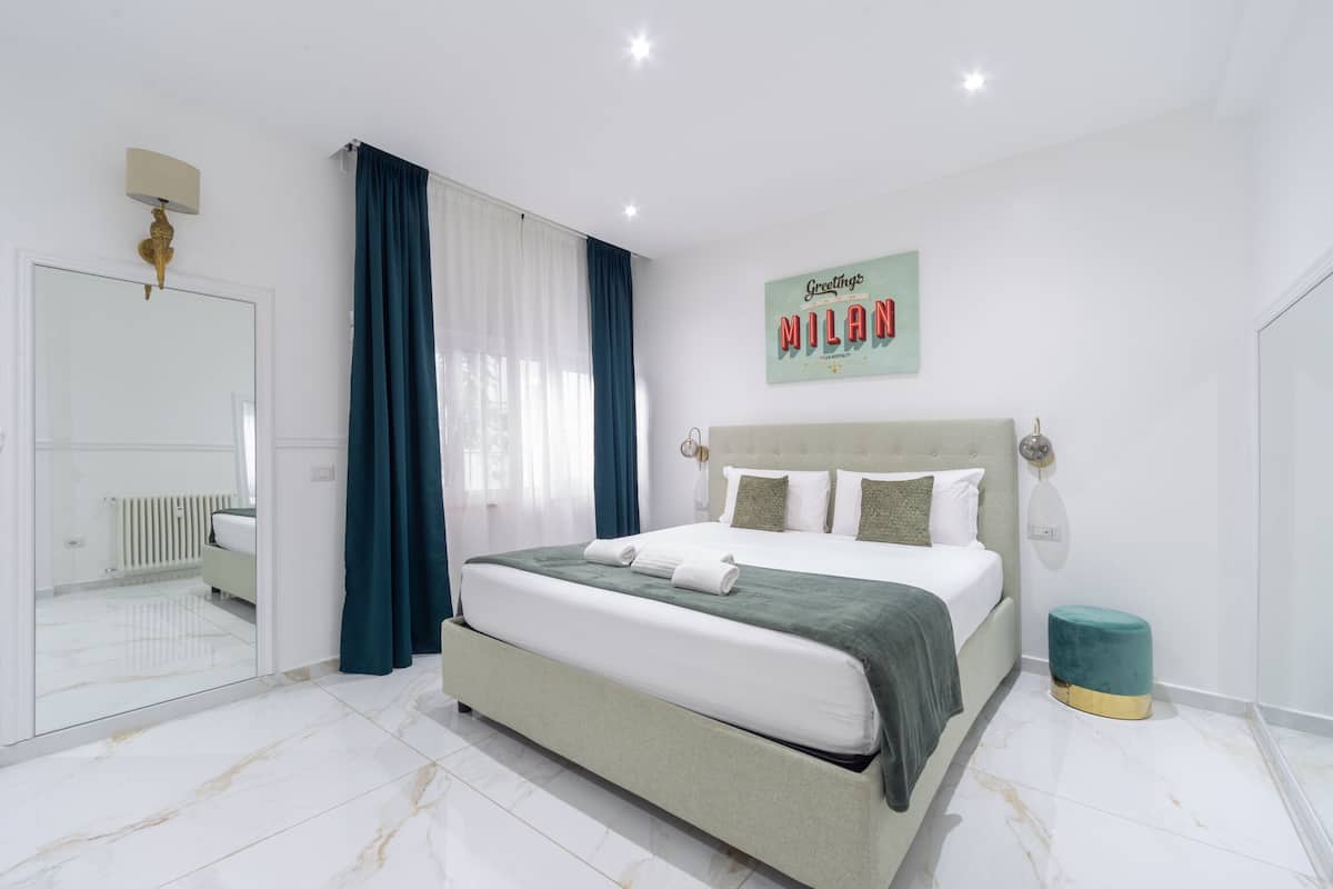 A modern bedroom with a large bed, a mirror, a wall clock, and a 'Greetings Milan' sign.