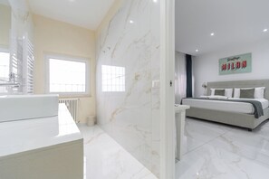 A modern bathroom with a white marble countertop and sink, a large window, and a bed with a grey headboard in the adjacent room.