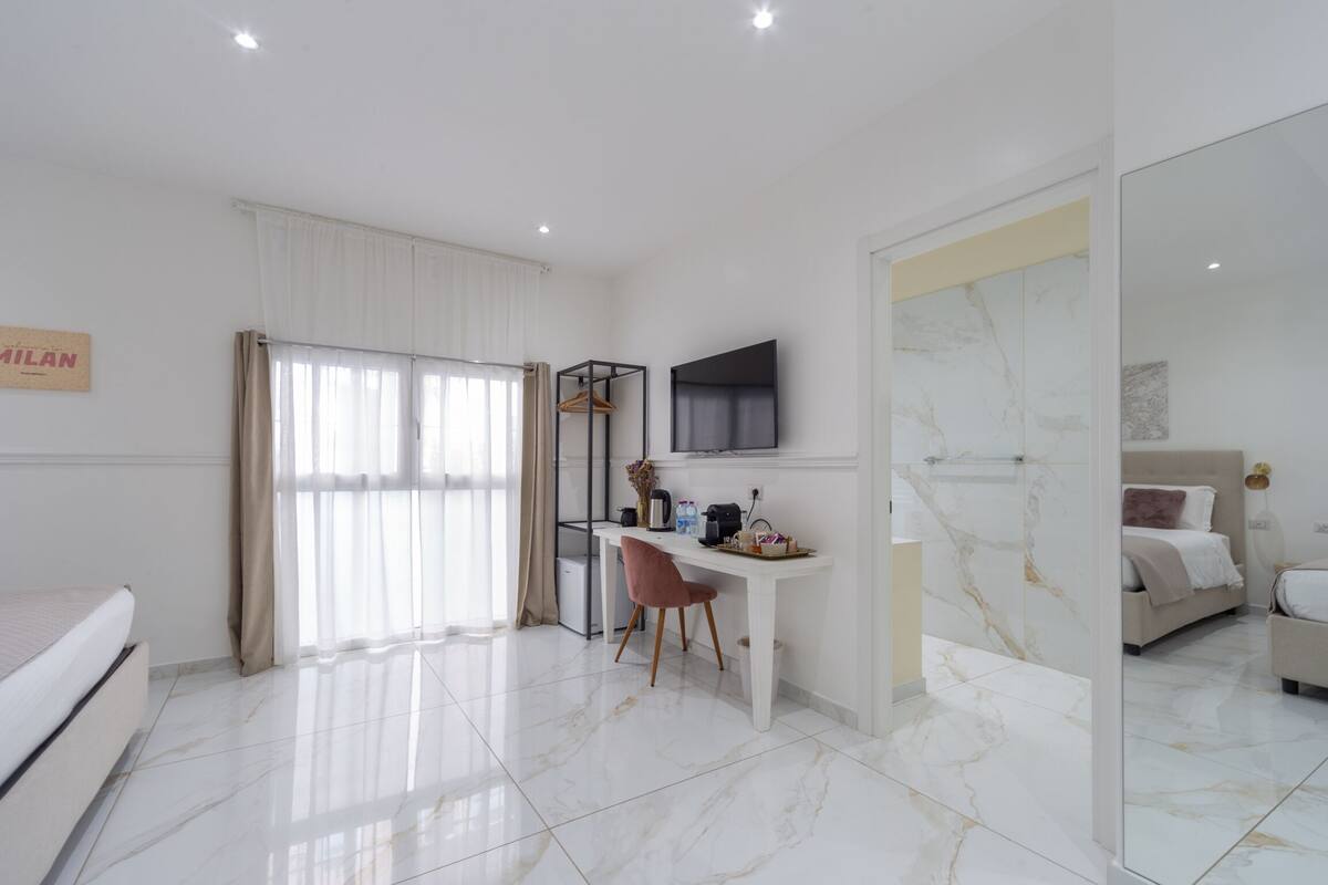 A modern hotel room with a bed, a desk with a chair, a TV mounted on the wall, and a bathroom with a mirror and sink.
