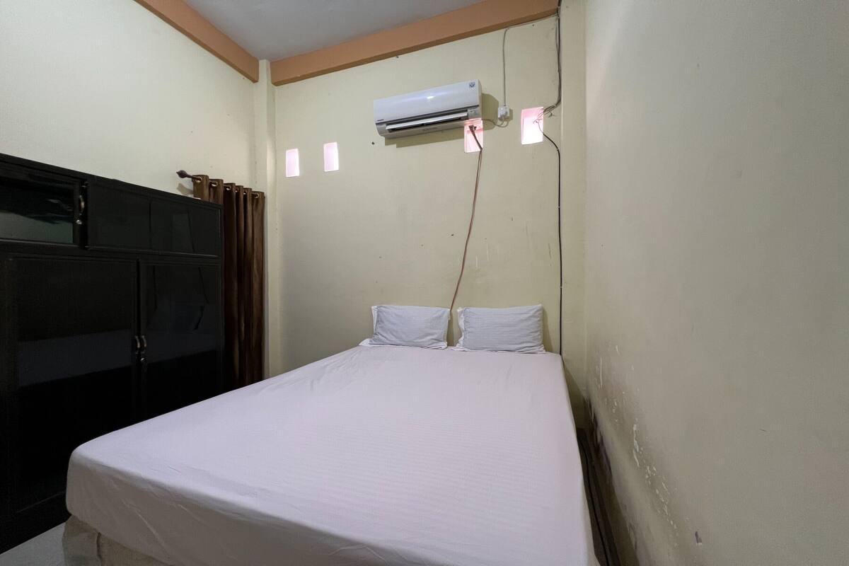 Un lit simple avec un matelas et des oreillers blancs, une armoire de couleur foncée et un climatiseur fixé au mur.