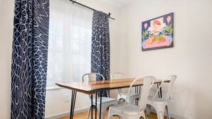 A dining area with a wooden table and white chairs, a window with patterned curtains, and a colorful painting on the wall. - Dog-friendly Single-Family Home w Free Parking, Washer, Dryer in NYC "Nantucket" (The Bronx)