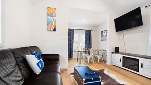 A living room with a leather sofa, a blue and white pillow, a wooden coffee table, a white cabinet with a TV, and a dining area with white chairs and a table. - Dog-friendly Single-Family Home w Free Parking, Washer, Dryer in NYC "Nantucket" (The Bronx)