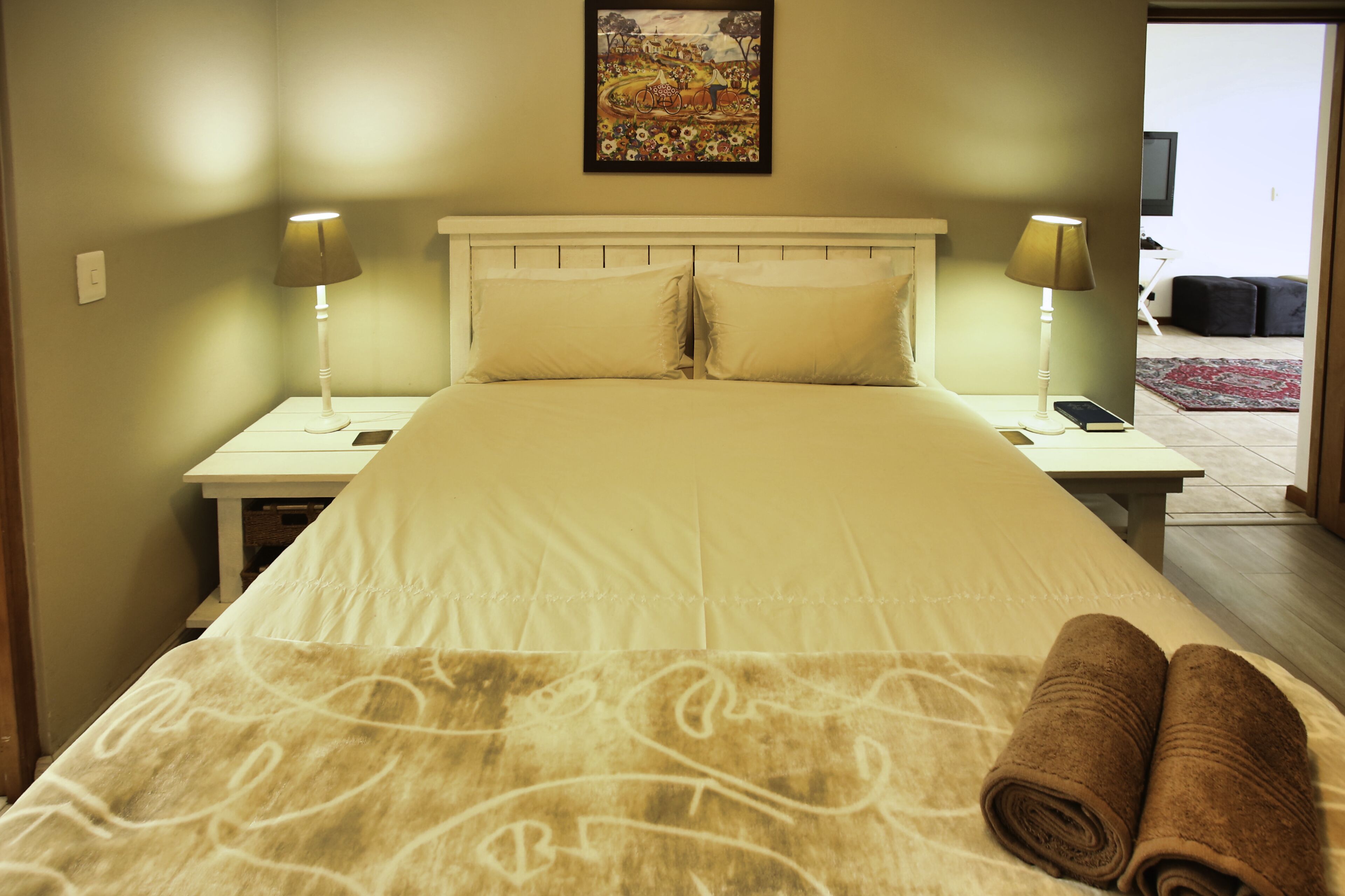 A hotel room with a large bed, two bedside tables, a nightstand, a painting on the wall, and a view of the outside.