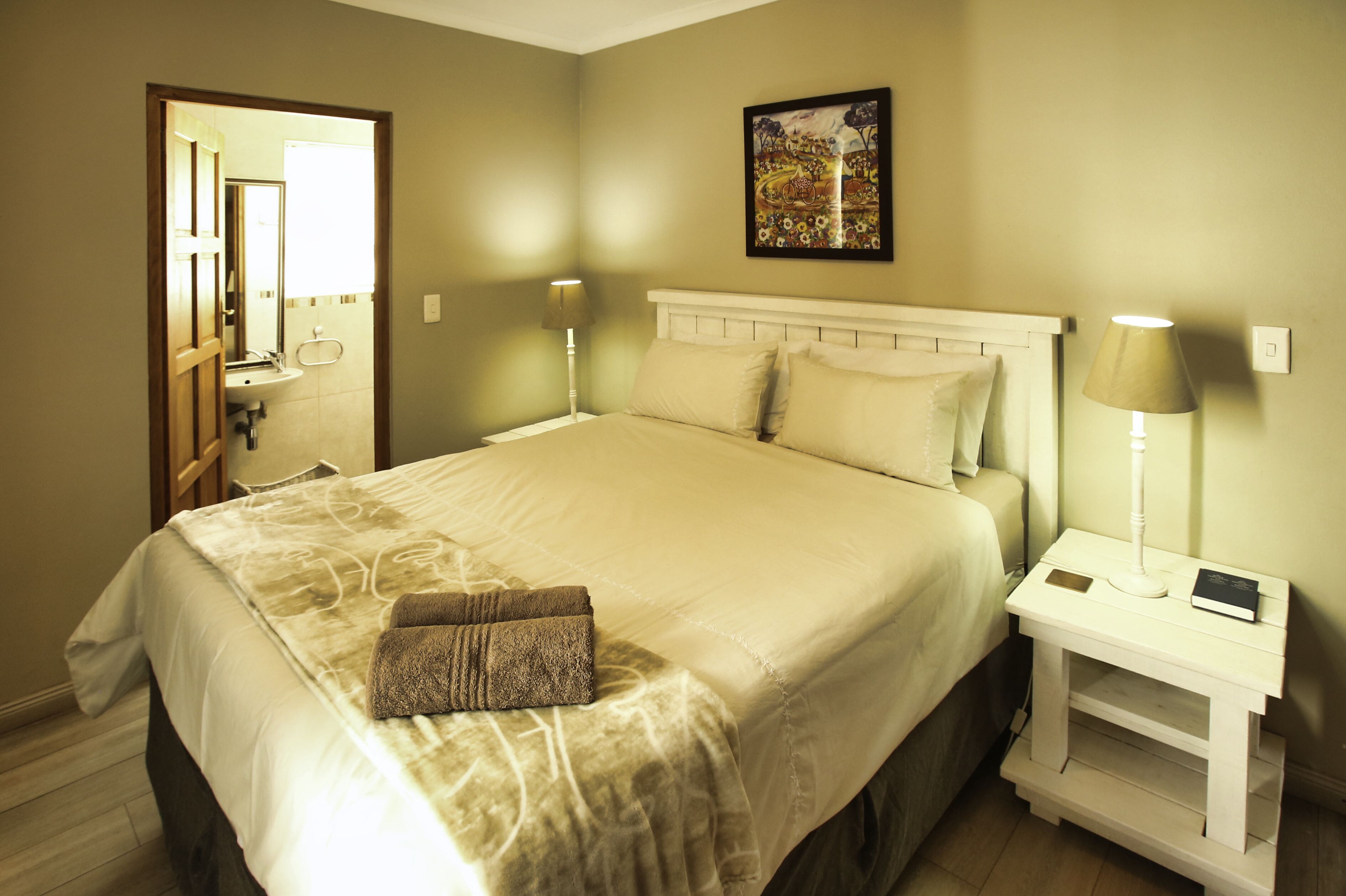 Una cama bien hecha con edredón color beige y dos almohadas, una mesita de noche con lámpara y un cuadro en la pared.