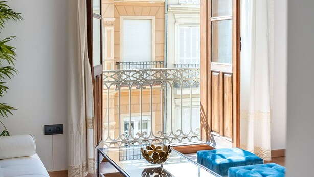 Un salon moderne avec une table basse en verre, des poufs bleus, un canapé blanc et une vue sur un immeuble à travers une porte vitrée.