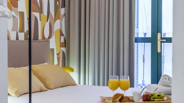 Ein Hotelzimmer mit einem Bett, zwei Orangensäften, einer Tablett mit Croissants und Obst sowie Blick ins Freie durch ein Fenster mit Vorhängen.