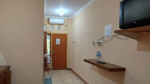 A hallway with a wooden door, a mounted television, a water dispenser, and a small shelf with a remote control. - Hotel O Rasya Homestay Near SD Negeri 6 Mataram (Mataram)