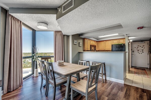 A dining area with a wooden table and chairs, a view through large windows, and a kitchen with wooden cabinets and appliances.
