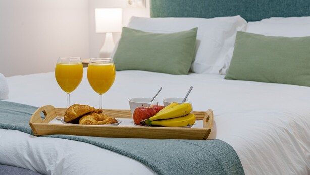 Un plateau de petit-déjeuner joliment préparé, avec des croissants, des bananes, des pommes et du jus d’orange, posé sur un lit recouvert de draps blancs et agrémenté d’oreillers verts.