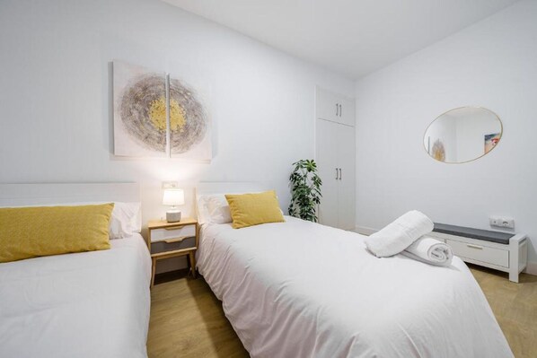 A hotel room with two beds, a bedside table with a lamp, a mirror, a plant, and a wardrobe.