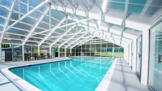A swimming pool with a glass ceiling structure, surrounded by chairs and a view of greenery outside. - Adirondack Retreat Inn & Suites - Superior Rental House (Lake George)