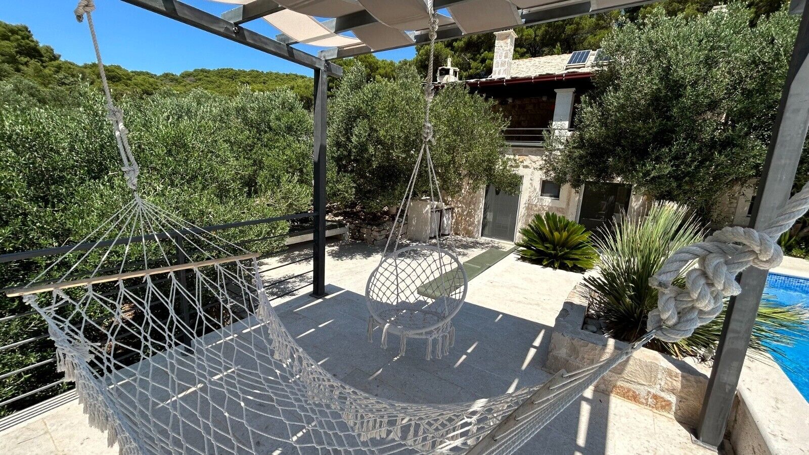 Eine Hängematze, die an einem Metallgestell befestigt ist, ein Stuhl und ein Pool in einer begrünten Außenanlage.