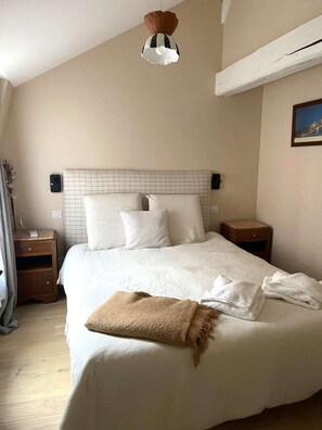 Ein ordentlich bezogenes Bett mit weißen Laken und einer braunen Decke, ein Nachttisch mit einer Lampe und einer Wandleuchte.