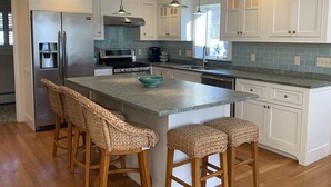 Une cuisine moderne avec des meubles blancs, un réfrigérateur en acier inoxydable, un four encastré et un plan de travail en granit. On y trouve quatre tabourets en osier et un grand îlot central avec un saladier.