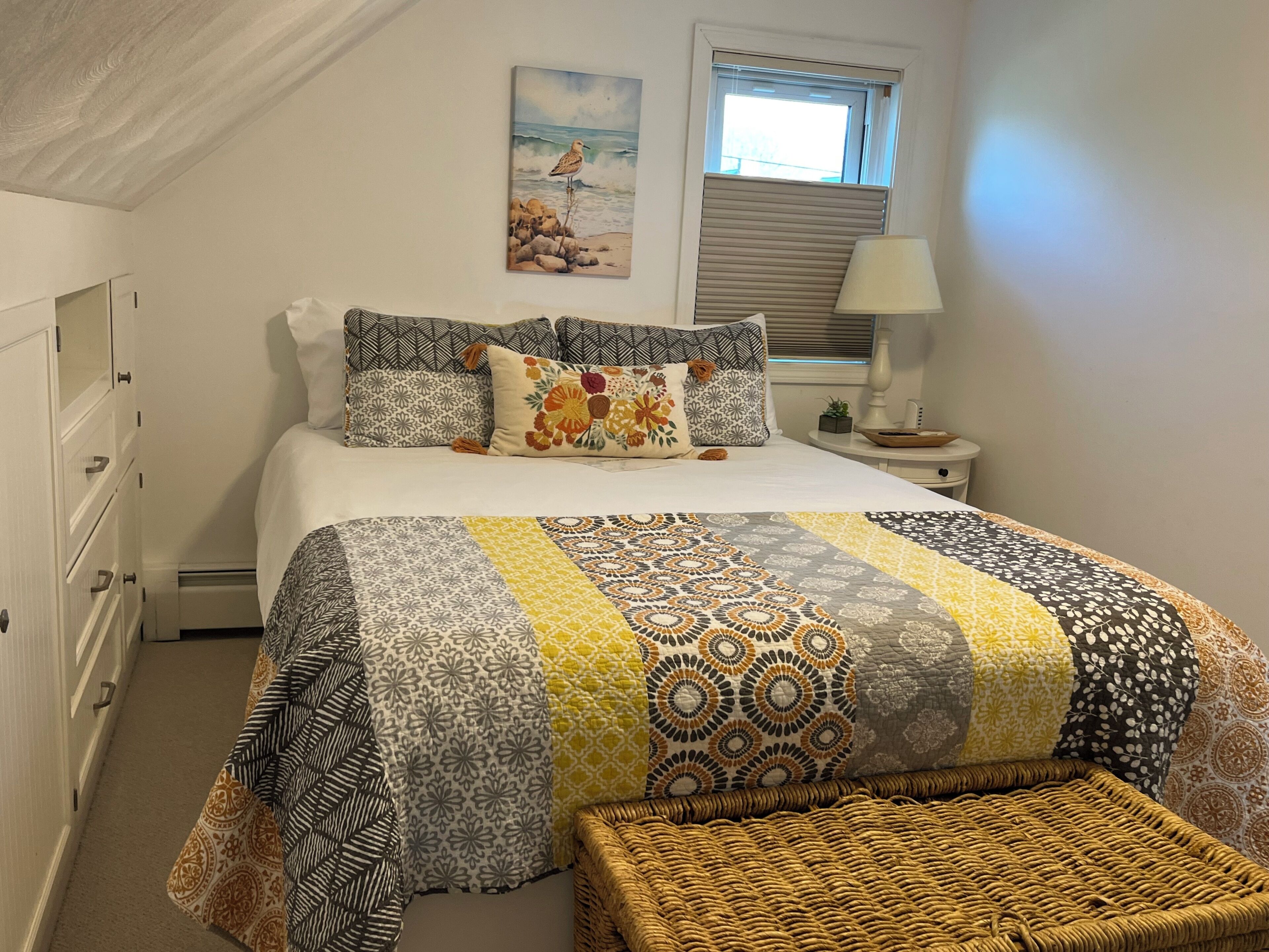 Un dormitorio con cama, almohadas, mesita de noche, lámpara, un cuadro y una ventana.
