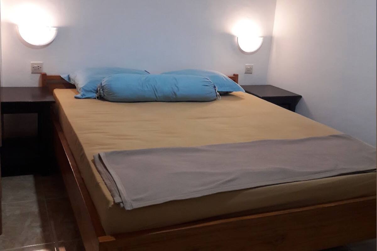 Een eenpersoonsbed met een beige matras en blauwe kussens, met aan beide zijden nachtkastjes met wandlampen.