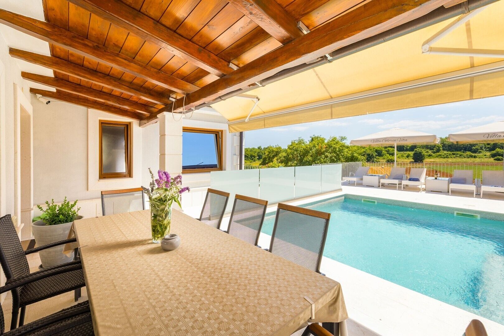 An outdoor pool area with a table and chairs, a wooden ceiling, and a view of the landscape.