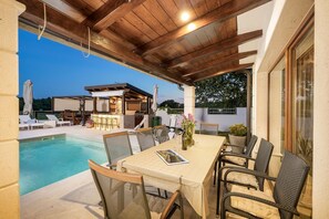 Ein Außenpoolbereich mit überdachter Terrasse, Liegestühlen und einem Esstisch mit Stühlen.