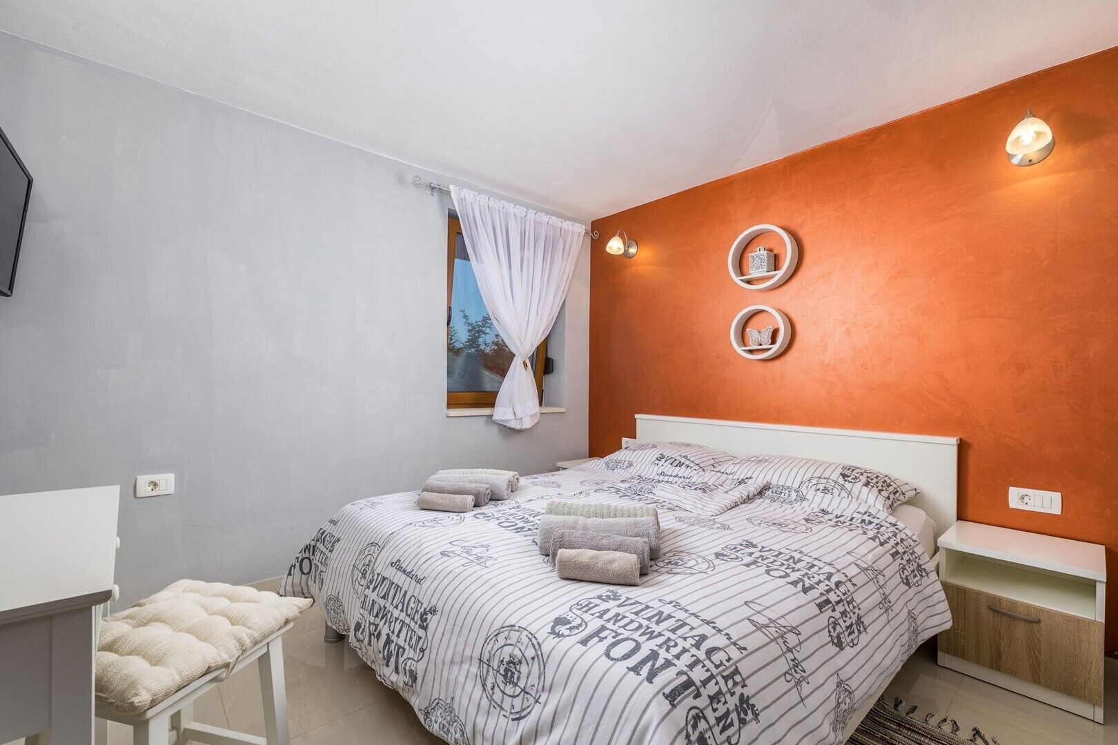 A bedroom with a bed, a bedside table, a chair, a window with curtains, and wall-mounted lights.