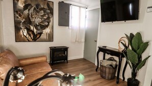 Ein Wohnzimmer mit einer Couch, einem Gemälde, einem Fernseher, einem kleinen Tisch, einer Pflanze und einem Korb.