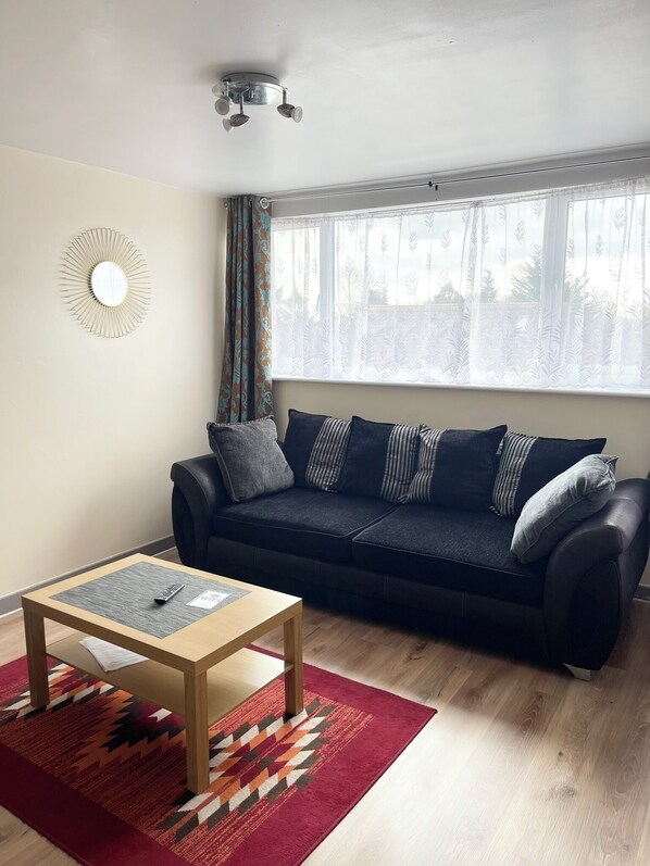 A living room with a dark sofa, a wooden coffee table, a patterned rug, and a window with sheer curtains. - 
Penn Gardens - Two Bedroom Apartment (Romford, Greater London, United Kingdom)
