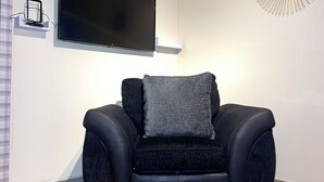A modern living room with a black leather sofa, a mounted flat-screen TV, a round glass coffee table with a cactus centerpiece, and a decorative mirror on the wall. - 
Penn Gardens - Two Bedroom Apartment (Romford, Greater London, United Kingdom)
