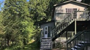 A two-story building with a staircase leading to a deck, surrounded by trees and a grassy area with wooden logs.